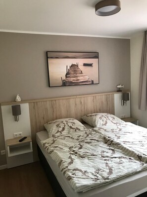 A double bed with a patterned bedspread, a wooden headboard, a wall-mounted picture of a pier, bedside lamps, and a small shelf with a remote control. - Modernes und Helles 2-zimmer-apartment in Ruhiger Lage im Haus 5/3 Hund Erlaubt - Modernes und (Plau am See)