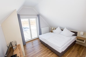 A bedroom with a sloped ceiling, a large bed with white bedding, a wooden floor, a nightstand with a lamp, and a view through a sliding door to the outside.