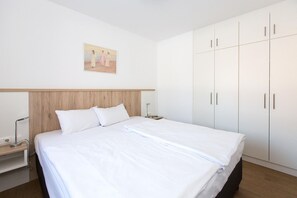 A neatly made bed with white linens, a wooden headboard, and a nightstand with a lamp. There is a large wardrobe with multiple doors and a painting on the wall above the bed.
