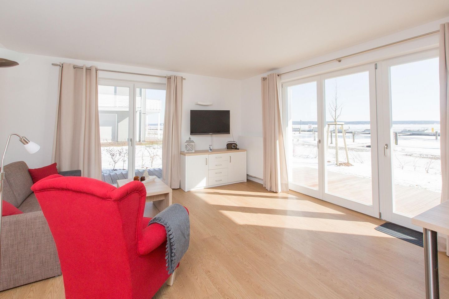Ein modernes Wohnzimmer mit einem roten Sofa, einem Fernseher auf einem weißen Schrank und großen Schiebegläsern, die zu einem verschneiten Außenbereich führen.