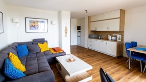 Un salon moderne avec un canapé gris, des coussins bleus et jaunes, une table basse blanche et un coin cuisine doté de meubles en bois et d’une table à manger.