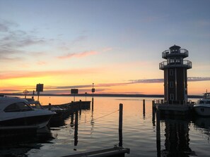 Un faro en un muelle con barcos, cielo despejado y un atardecer.