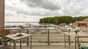A terrace with tables and chairs, overlooking a marina with boats and a forested hill in the background.