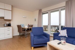 Un salon moderne avec un canapé bleu, une petite table avec une télécommande et un verre d’eau. On y trouve également un coin repas avec une table et des chaises, ainsi qu’une cuisine équipée de placards et d’appareils électroménagers.