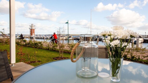 A glass table with a clear pitcher and a vase of white flowers, overlooking a marina with boats and a cyclist in the background.
