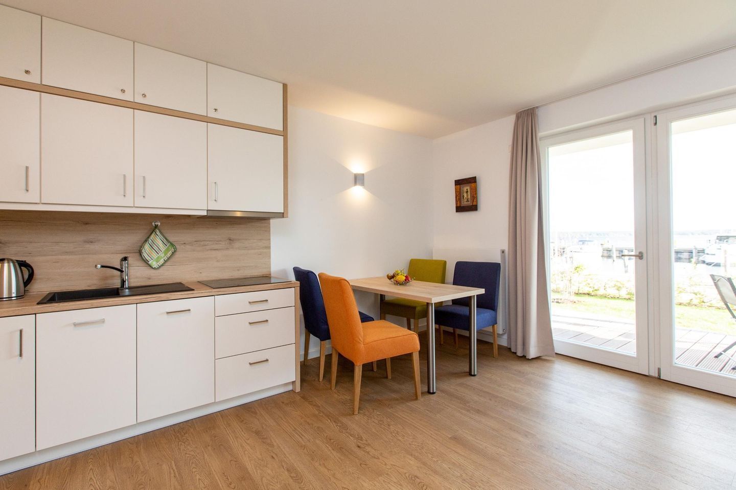 Een moderne keuken met witte kastjes, een roestvrijstalen waterkoker en een houten aanrecht. Er is een eethoek met een tafel en kleurrijke stoelen.