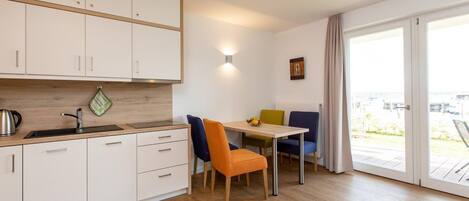 Een moderne keuken met witte kastjes, een roestvrijstalen waterkoker en een houten aanrecht. Er is een eethoek met een tafel en kleurrijke stoelen.