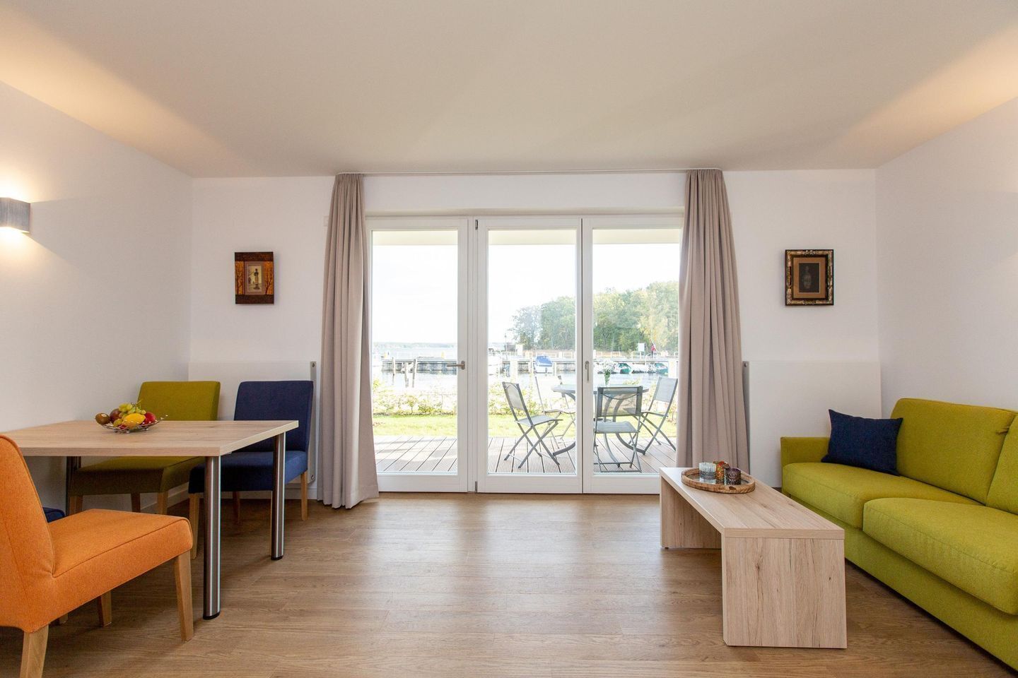 Una sala moderna con piso de madera, una mesa de comedor con sillas, un sofá y una pequeña mesa de centro. Hay una puerta corrediza de vidrio que da a un espacio exterior con sillas y una mesa.