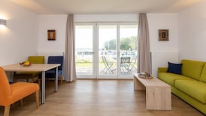 A modern living room with a wooden floor, a dining table with chairs, a sofa, and a small coffee table. There is a sliding glass door leading to an outdoor area with chairs and a table.