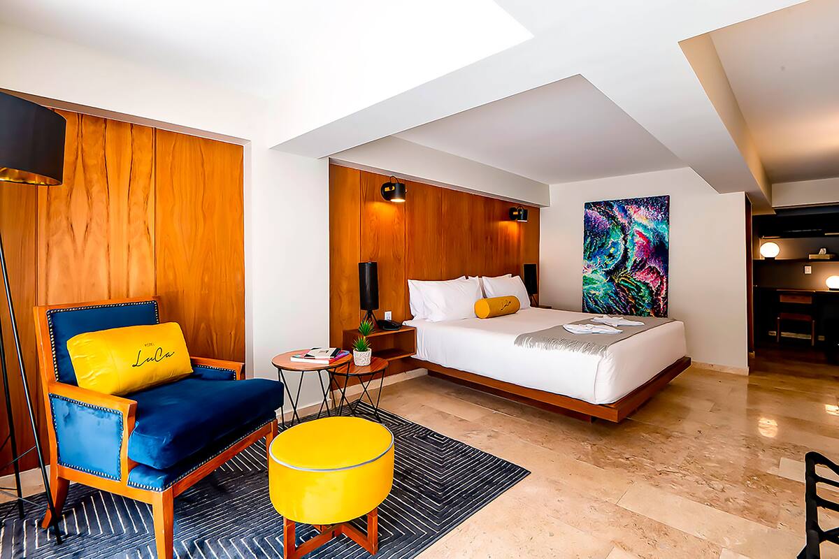 A modern hotel room with a bed, a blue armchair, a yellow ottoman, and a wooden accent wall.