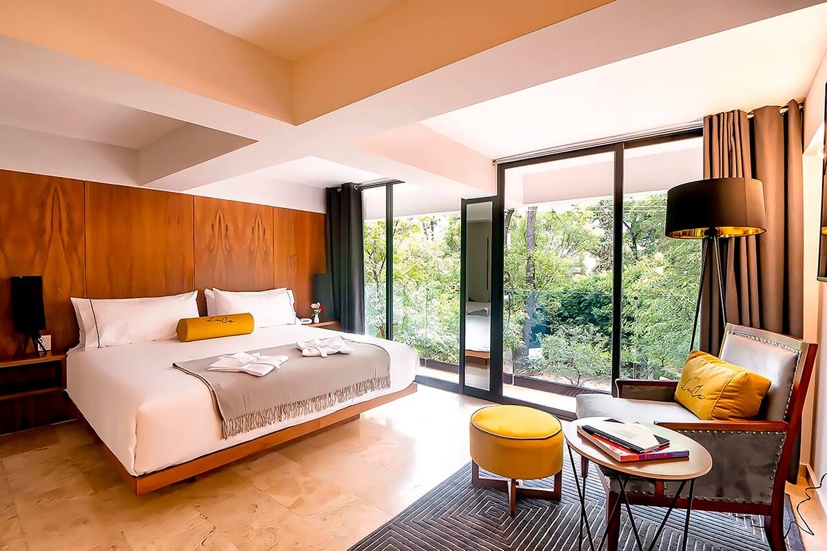 A modern hotel room with a large bed, a seating area with a chair and ottoman, and a balcony with a view of greenery.