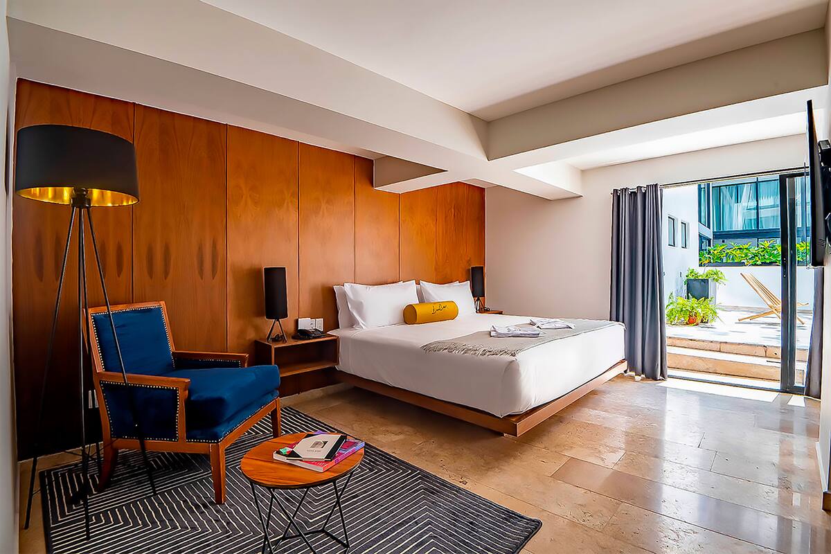 A modern hotel room with a large bed, a blue armchair, a bedside table with a lamp, and a view of the outdoors.