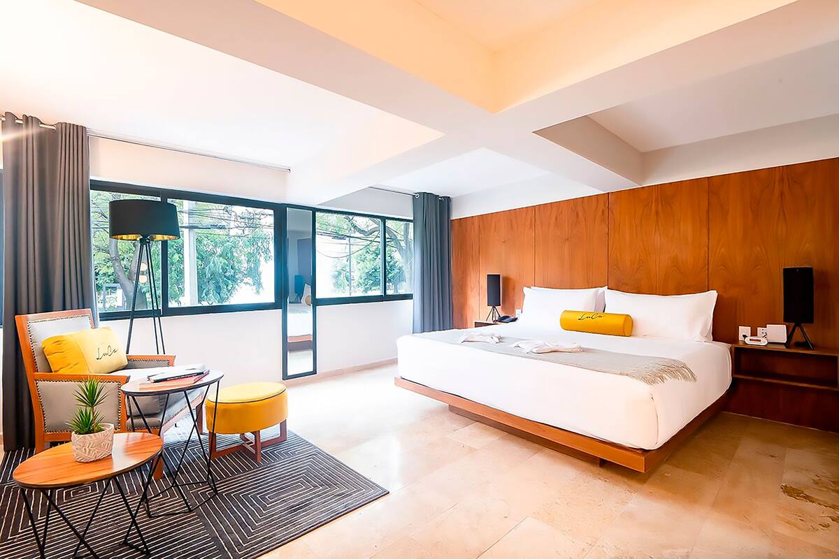 A modern hotel room with a large bed, a sitting area with a chair and table, and a view through large windows.