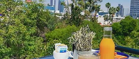 A breakfast setup with a coffee cup, orange juice, and a plate of food on a table with a city view in the background.