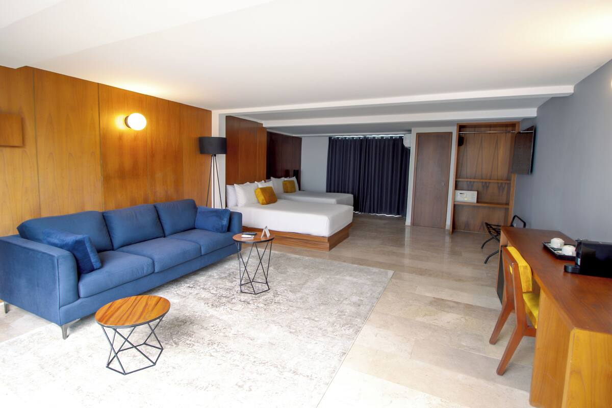 A modern hotel room with a blue sofa, a wooden dining table, a small round coffee table, a bed with white bedding, and a wooden wardrobe.