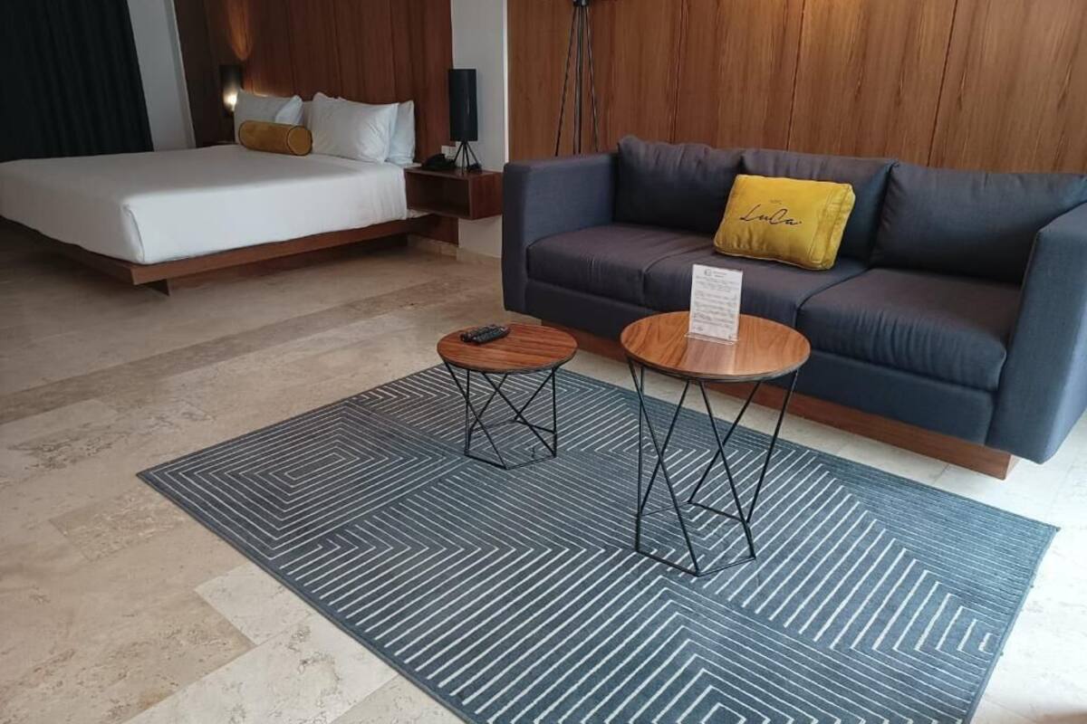 A modern hotel room with a bed, a sofa, two small tables, and a patterned rug.