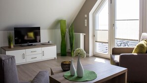 A modern living room with a flat-screen TV, a sofa, a coffee table, and a large window with blinds.