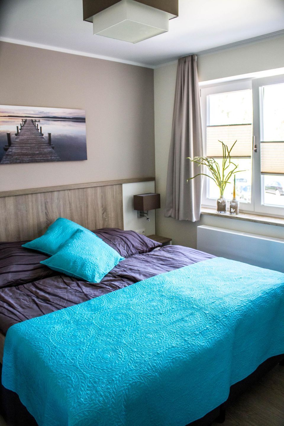 A hotel room with a bed, a nightstand, a lamp, a window with curtains, and a painting on the wall.