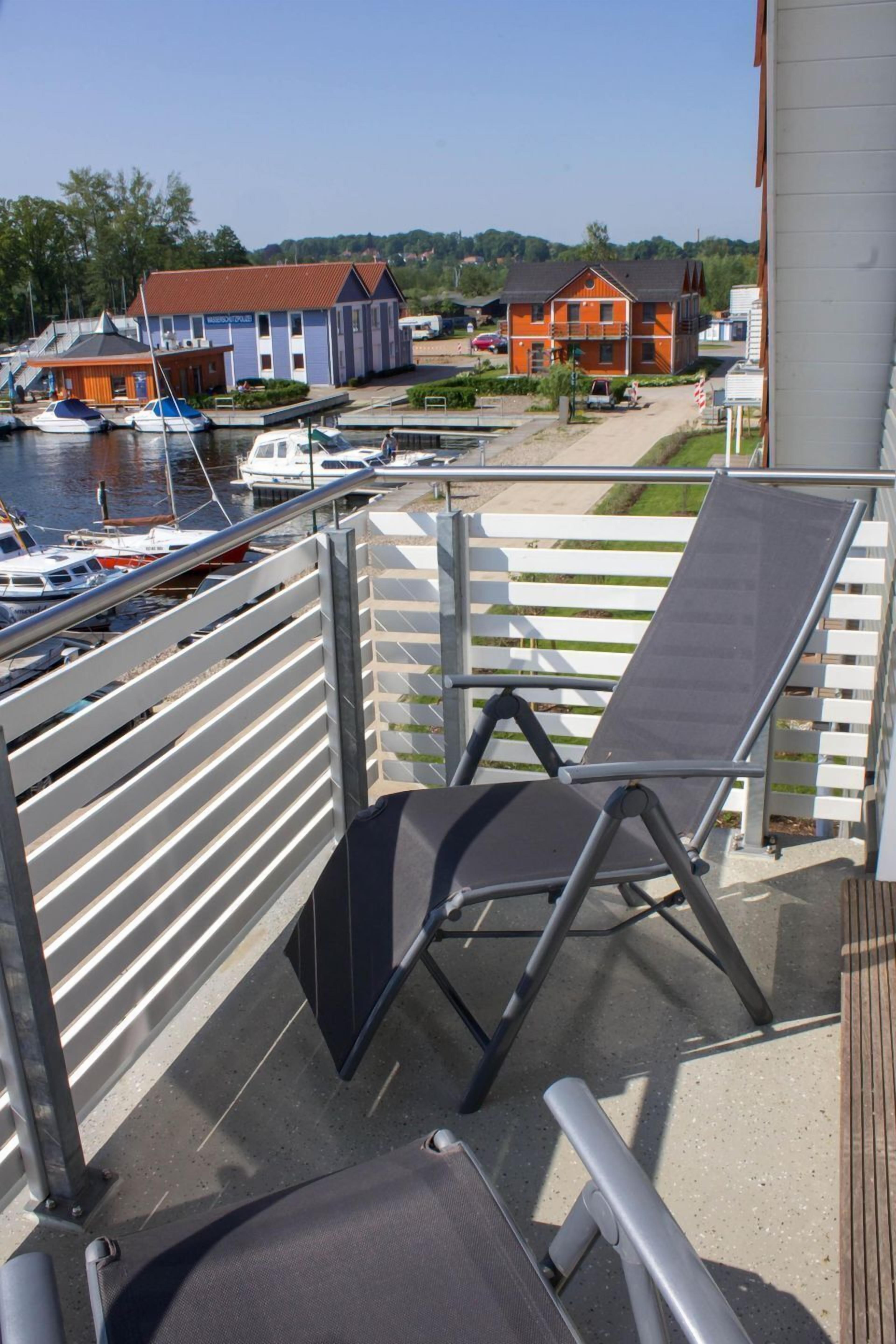 A balcony with a metal railing, a black lounge chair, and a view of boats and buildings in the background.