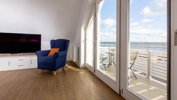 Un salon moderne avec une grande télévision, un fauteuil bleu et un balcon offrant une vue sur la mer.