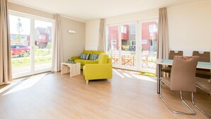 Un salón luminoso con un sofá amarillo, una mesa de comedor con sillas y grandes puertas correderas de vidrio que dan a un balcón.