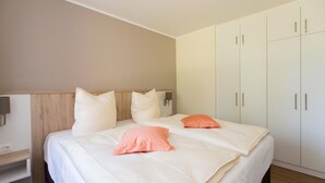 Habitación de hotel con dos camas, cabecero de madera, armario blanco y mesita de noche con lámpara.