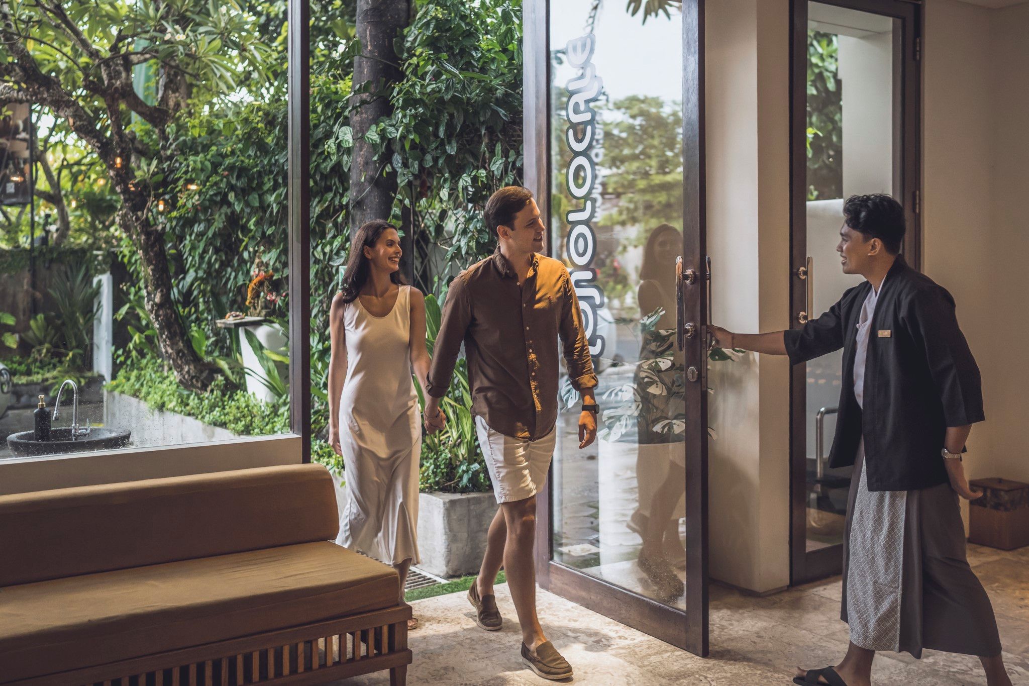 A hotel entrance with a glass door, a bench inside, and a view of greenery outside.