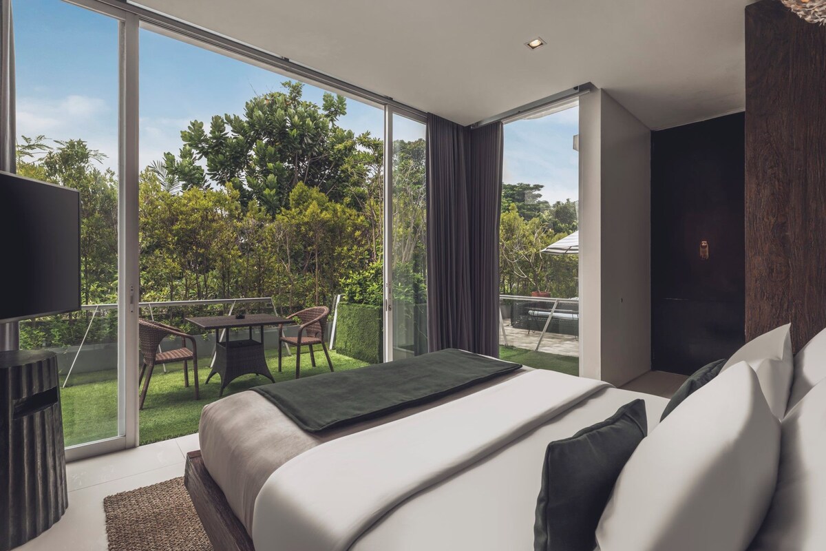 A modern hotel room with a large bed, a flat-screen TV, a balcony with a table and chairs, and a view of greenery.