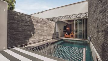 Un área de piscina moderna al aire libre con una pared de piedra, un espacio de descanso con cojines y un ventanal que da a una habitación.