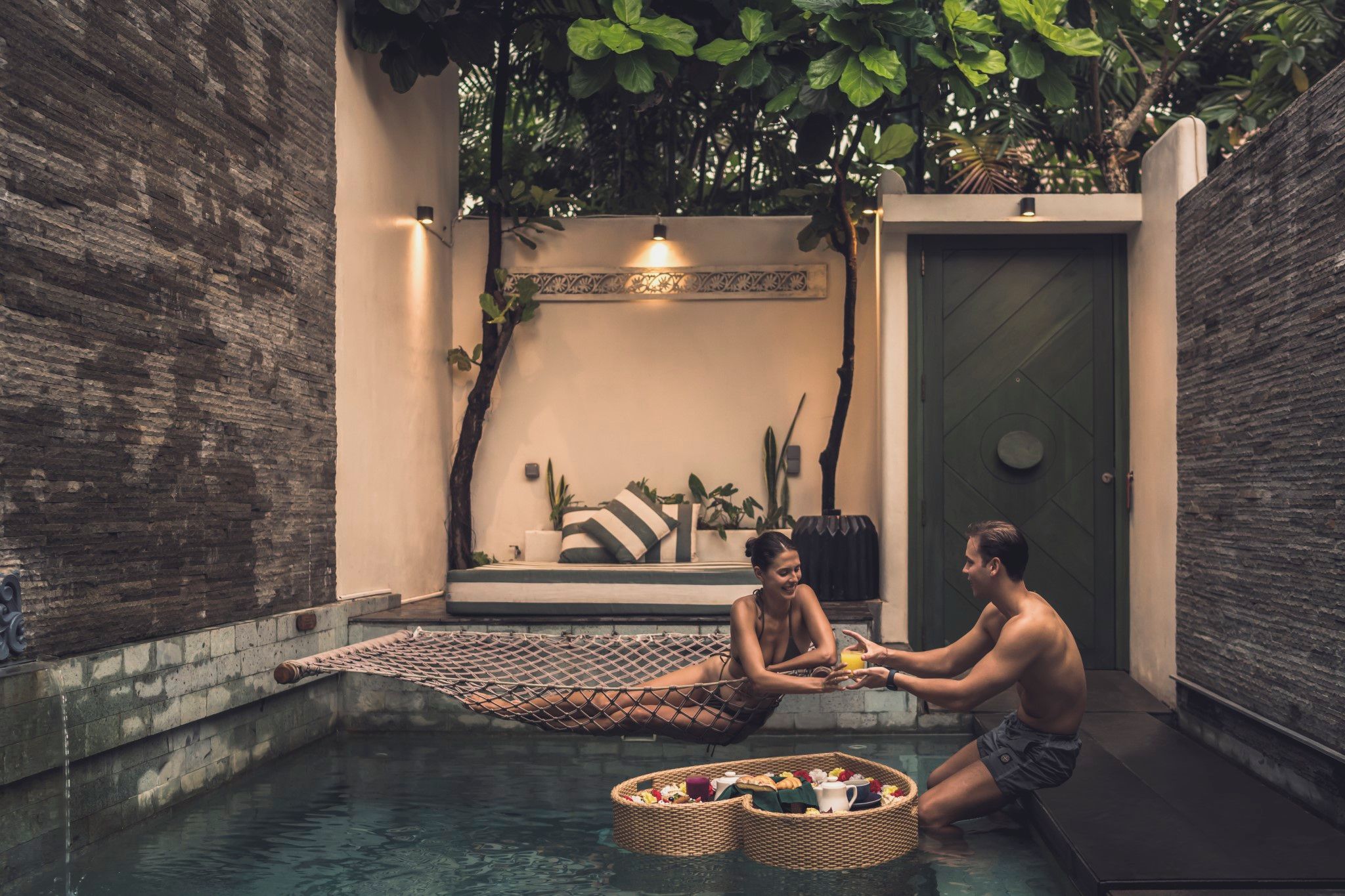 Una pareja descansando junto a una piscina con una hamaca y cestas de frutas y flores.