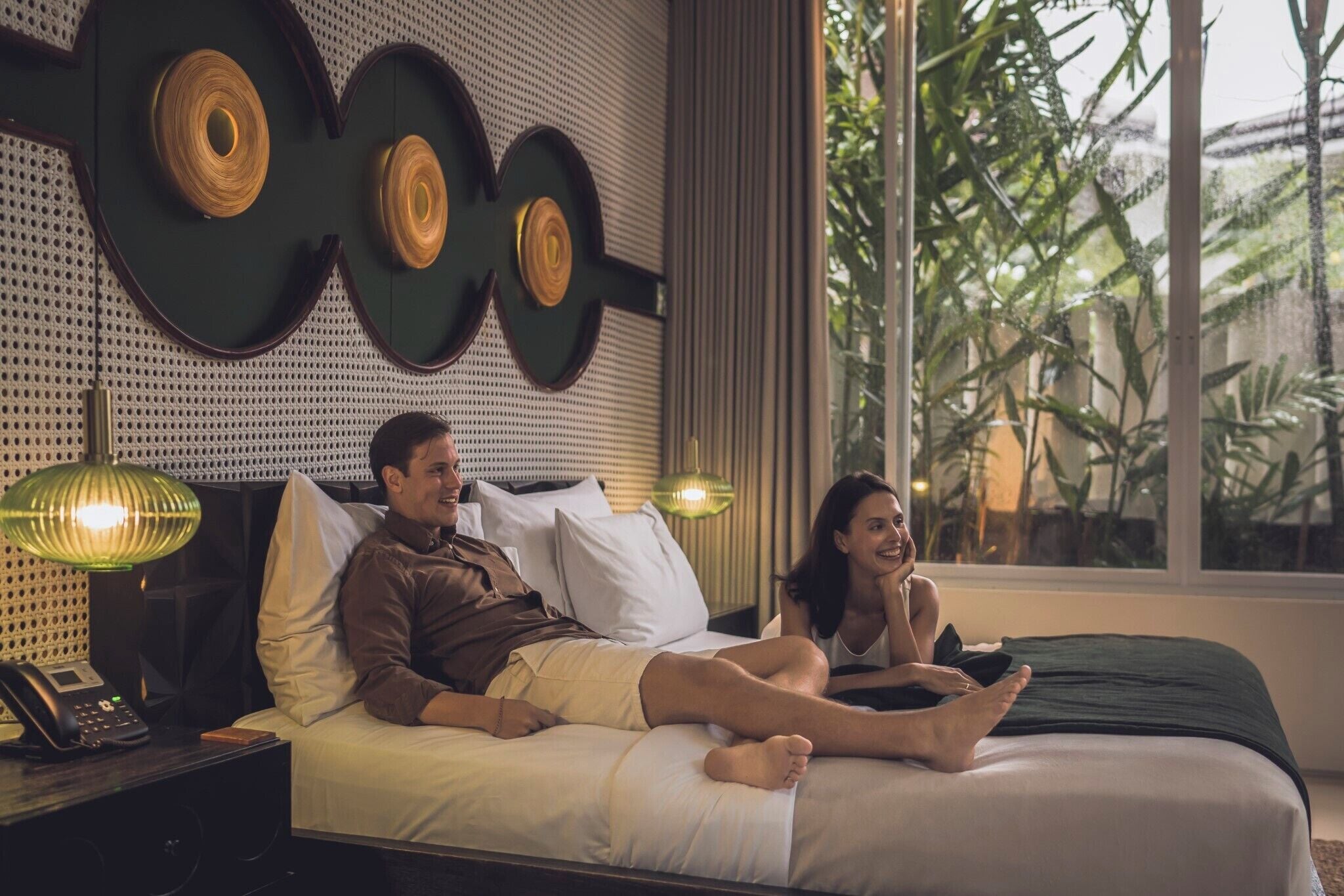 Quarto com cama, duas luminárias de cabeceira, mesa de cabeceira com telefone e janela com vista para área verde.