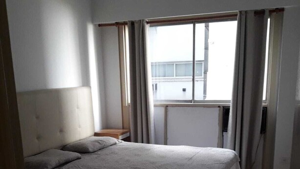 Un dormitorio con cama, mesita de noche, ventana con cortinas y vista a un edificio.