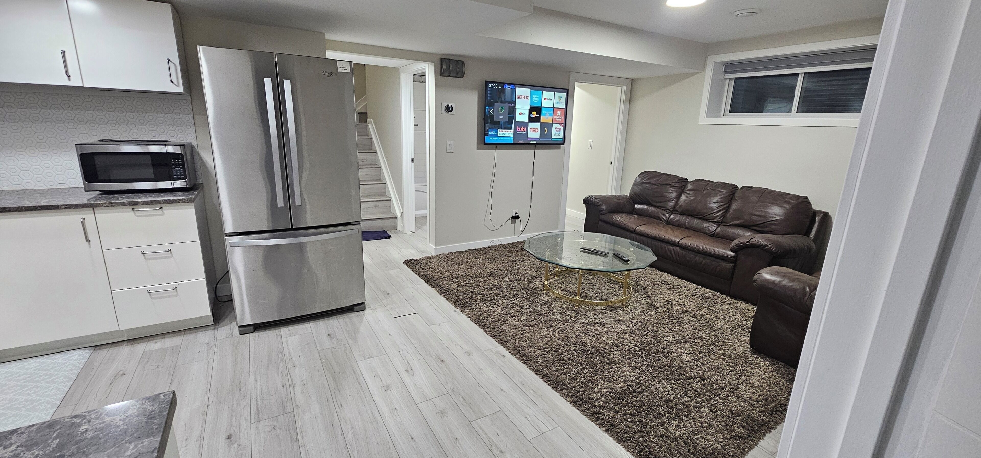 A compact living space with a kitchenette, a glass coffee table, a brown leather sofa, and a flat-screen TV mounted on the wall.
