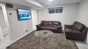 Un salon avec deux canapés en cuir marron, une table basse en verre, un téléviseur fixé au mur et un sol recouvert d’une moquette.