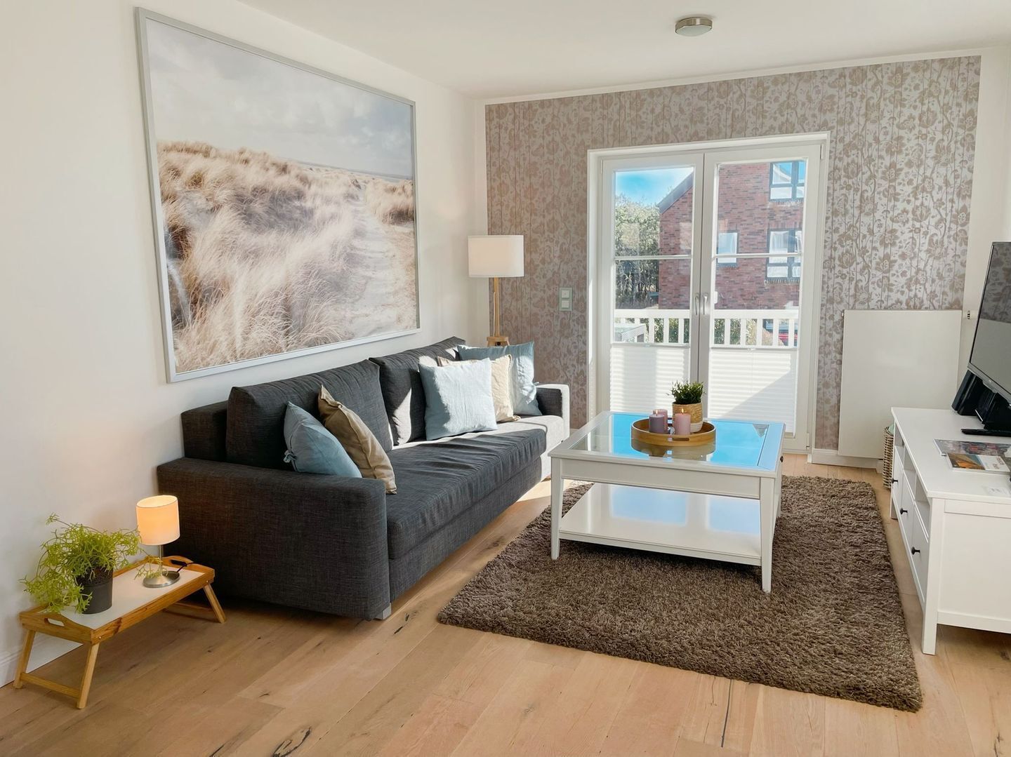 Ein modernes Wohnzimmer mit einem grauen Sofa, einem Glastisch, einer Lampe und einem Fernseher auf einer weißen Kommode.