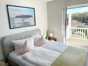 A bedroom with a bed, pillows, a bedside table with a lamp, a framed picture on the wall, and a balcony with a view of trees. - Sea breeze DUE/08 - dune bottom (Sylt)