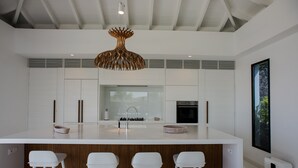 Una cocina moderna con una isla central, taburetes blancos de bar, una lámpara colgante original y un ventanal grande.