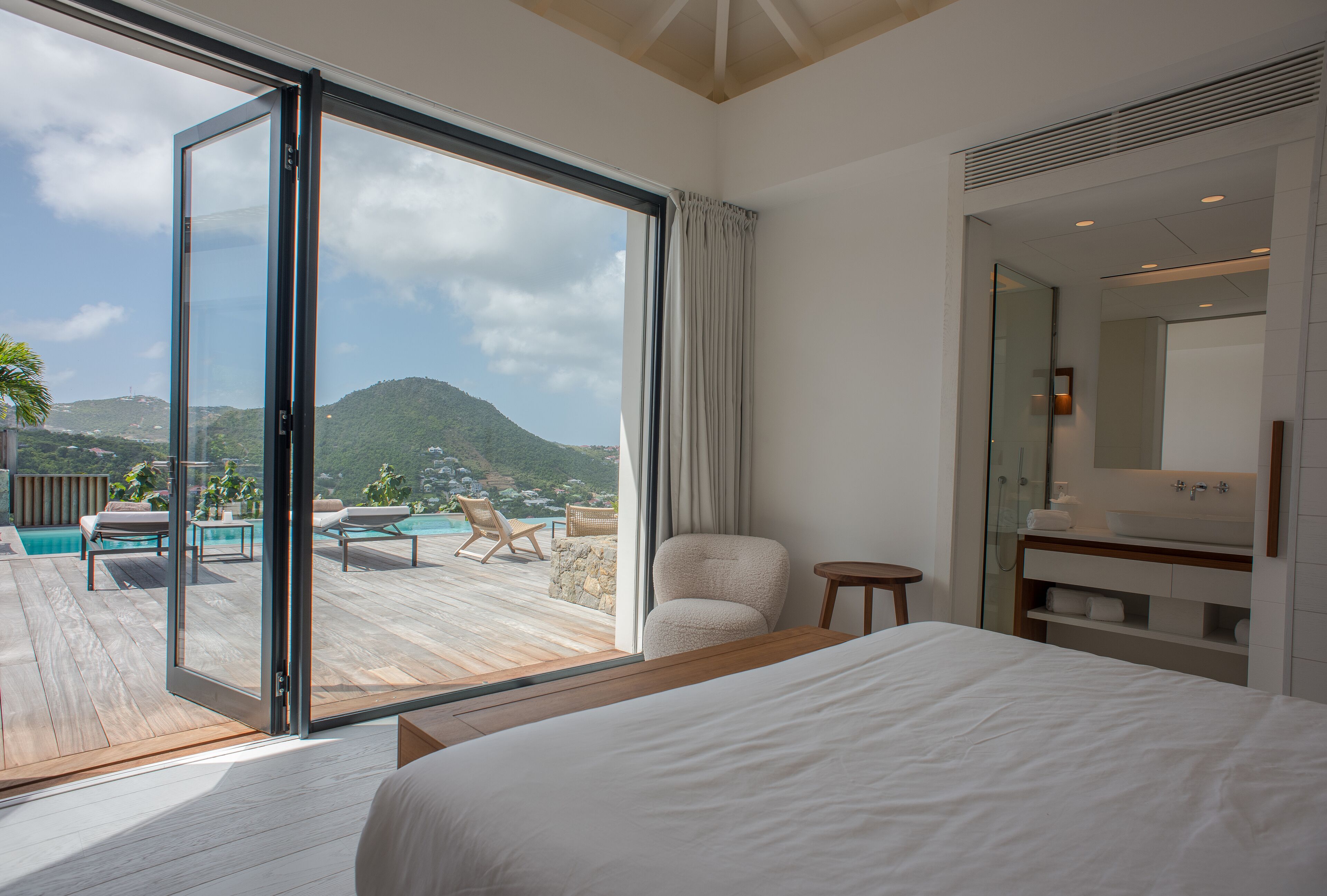 A bedroom with a large sliding glass door leading to a deck with a pool and chairs, a wooden bed, and a bathroom with a mirror and sink.