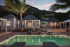 Un área junto a la piscina con sillones reclinables, una terraza de madera y una casa con porche cubierto.