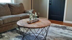 Una sala de estar con un sofá estampado, una mesa de centro redonda de madera con patas metálicas y una puerta al fondo.