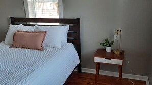 Una cama bien hecha con almohadas blancas y rosas, una mesita de noche con lámpara y una ventana con persianas.