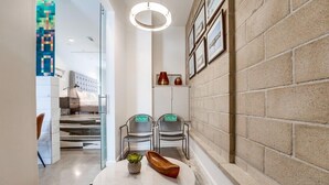 A modern hotel lobby with a marble floor, a round table with a plant and a wooden bowl, two chairs labeled "Little Room," and a wall with framed pictures and a brick accent wall.