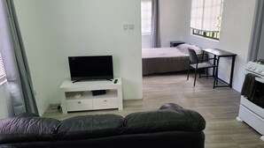 Uma sala de estar com televisão de tela plana em um suporte branco, um sofá de couro preto, uma mesa pequena com uma cadeira, uma cozinha compacta com fogão e uma janela com persianas.