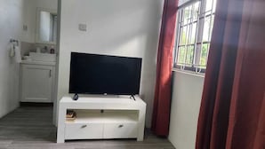 A room with a flat-screen TV on a white stand, a window with red curtains, and a white cabinet with a mirror. - Steps away from a beach & local transportation. Cozy one bedroom apartment (Saint George's)