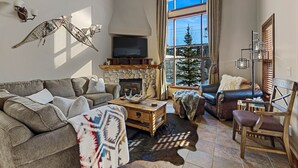 A cozy living room with a fireplace, a large window with a view of a snowy landscape, a leather sofa, and a wooden coffee table. - Bright, Ski-in/Ski-out Townhouse with A/C (Sun Peaks)
