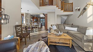 A spacious living area with a large sectional sofa, a wooden coffee table, and a leather chair. A staircase with wooden railings leads to an upper level. - Bright, Ski-in/Ski-out Townhouse with A/C (Sun Peaks)