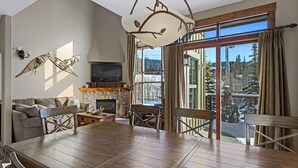 A spacious living room with a wooden dining table, comfortable seating, a fireplace, and a large window offering a view of a snowy landscape. - Bright, Ski-in/Ski-out Townhouse with A/C (Sun Peaks)