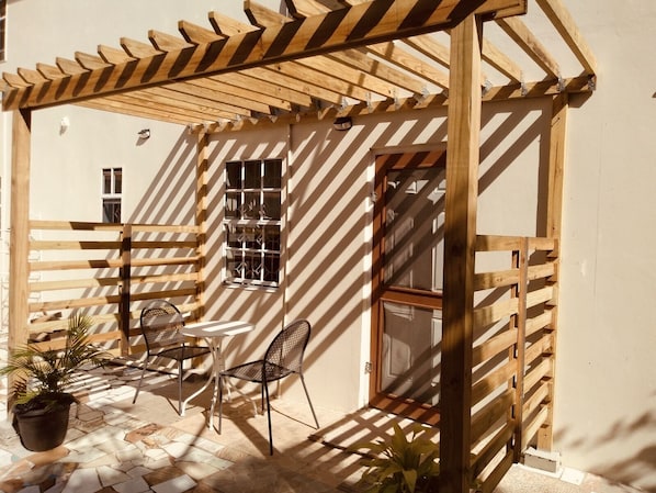 A covered patio area with a wooden pergola, a small table, two chairs, and potted plants.