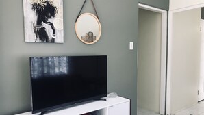 A modern living room with a flat-screen TV on a white cabinet, a framed artwork above the TV, and a round mirror on the wall. - Apartment - Walk to the Beach (Lucea)