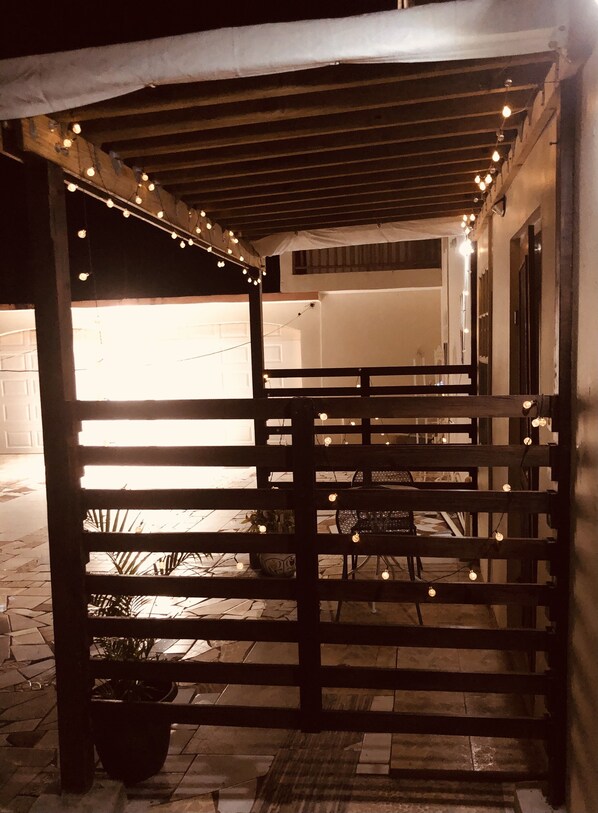 Un área de estar al aire libre techada, con luces colgantes, barandillas de madera y plantas en macetas.